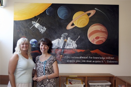 Teachers Karen Gronberg (left) and Julie Kennedy (right) stand before a mural in honor of a longtime board member and rocket designer